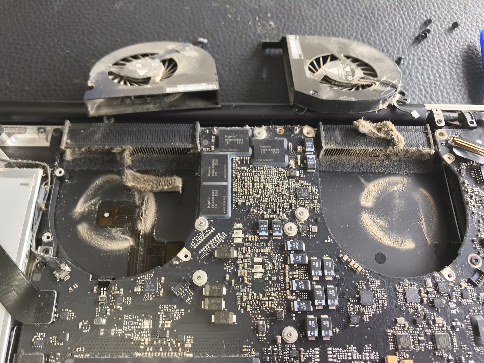 Interior de una MacBook Pro de 15 pulgadas con los dos ventiladores y los disipadores cubiertos de pelusa, y la marca de polvo dibujada en la placa