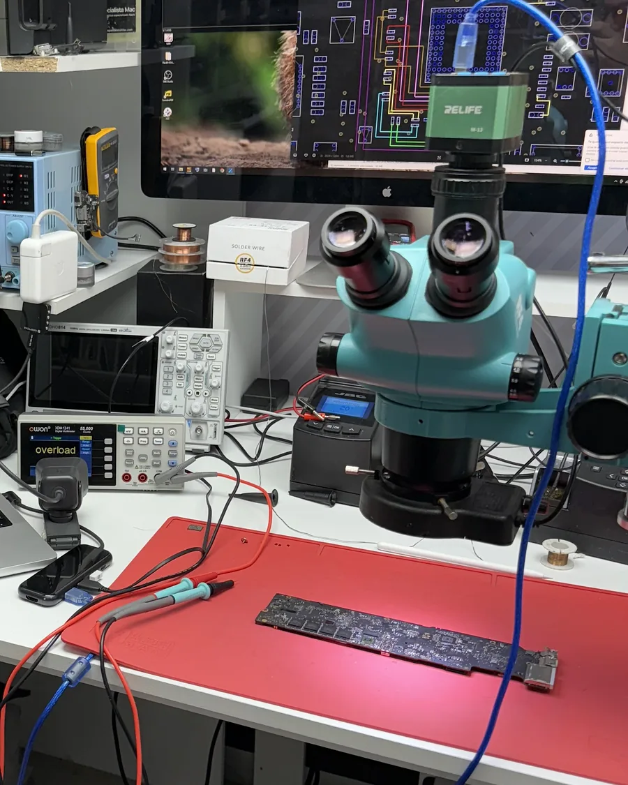 Banco del laboratorio con microscopio, multímetro de mesa y una placa lógica sobre el tapete rojo, con un boardview abierto en el monitor.
