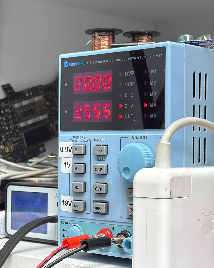 Fuente de poder regulable del laboratorio marcando 20,00 voltios y 3,555 amperios, con un cargador de Mac al lado.