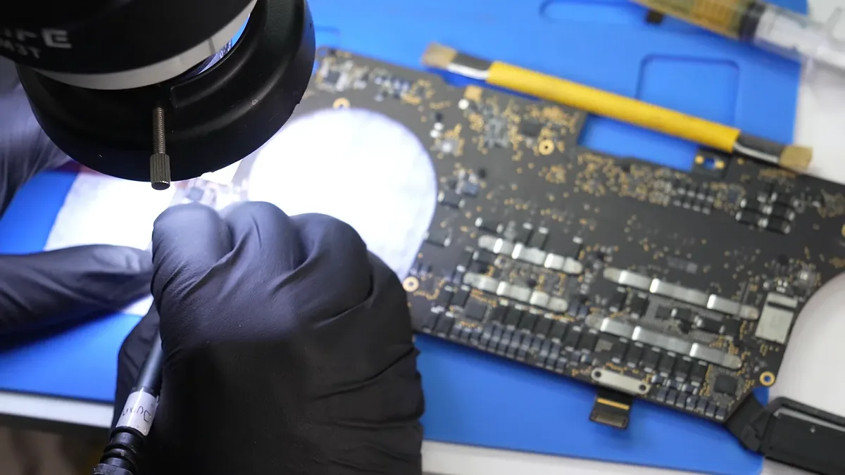 Mano con guante trabaja con el cautín bajo el lente del microscopio, junto a una placa de MacBook.