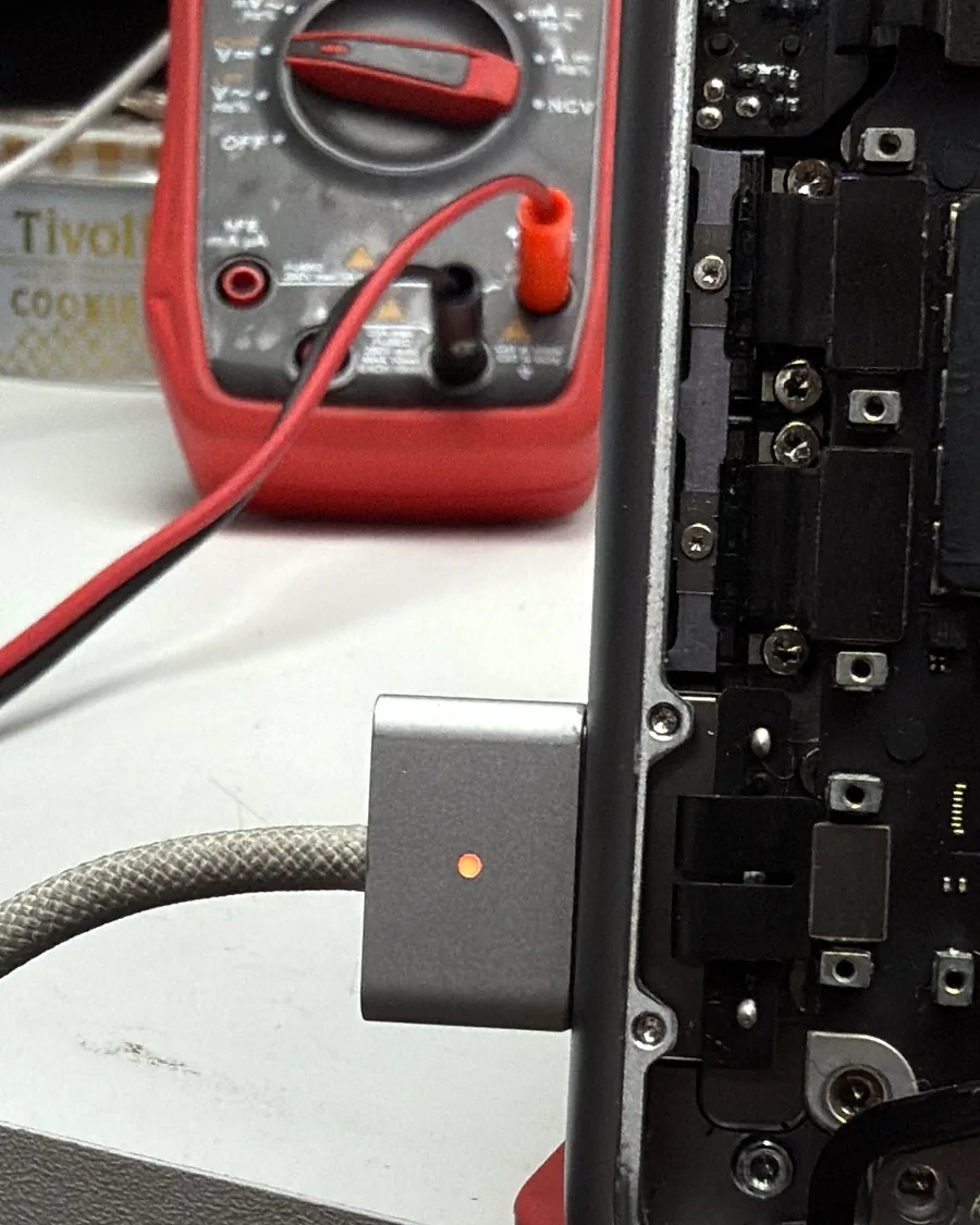 Conector MagSafe con la luz naranja encendida en una MacBook abierta, con un multímetro detrás.