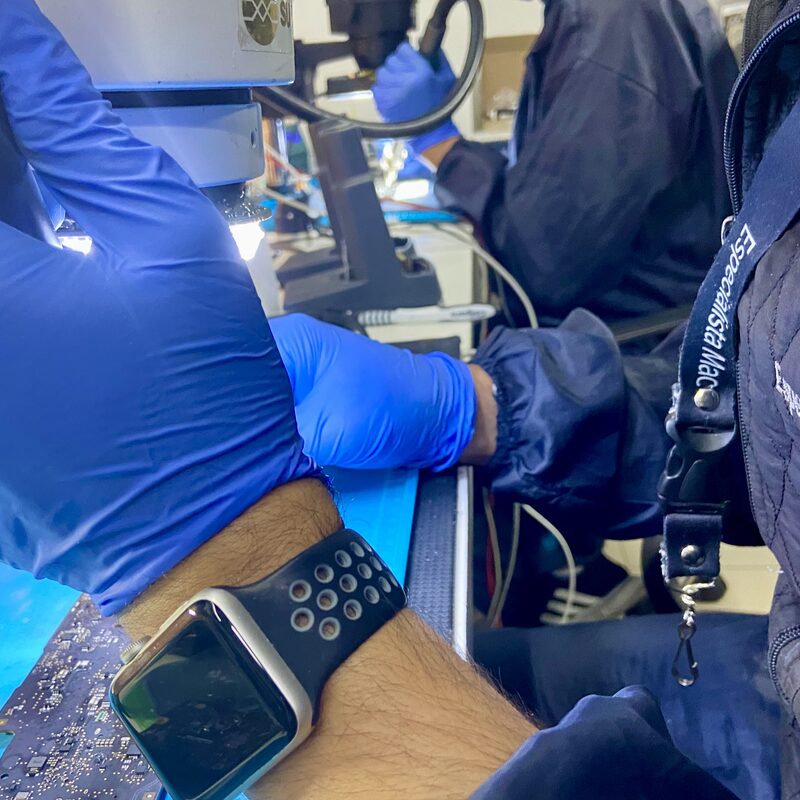 Técnico con guantes azules y credencial EspecialistaMac trabajando bajo microscopio en placa lógica